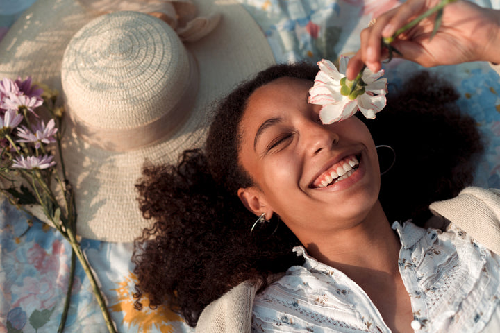 Girl holding white flower to her eye laughing on blanket with hat