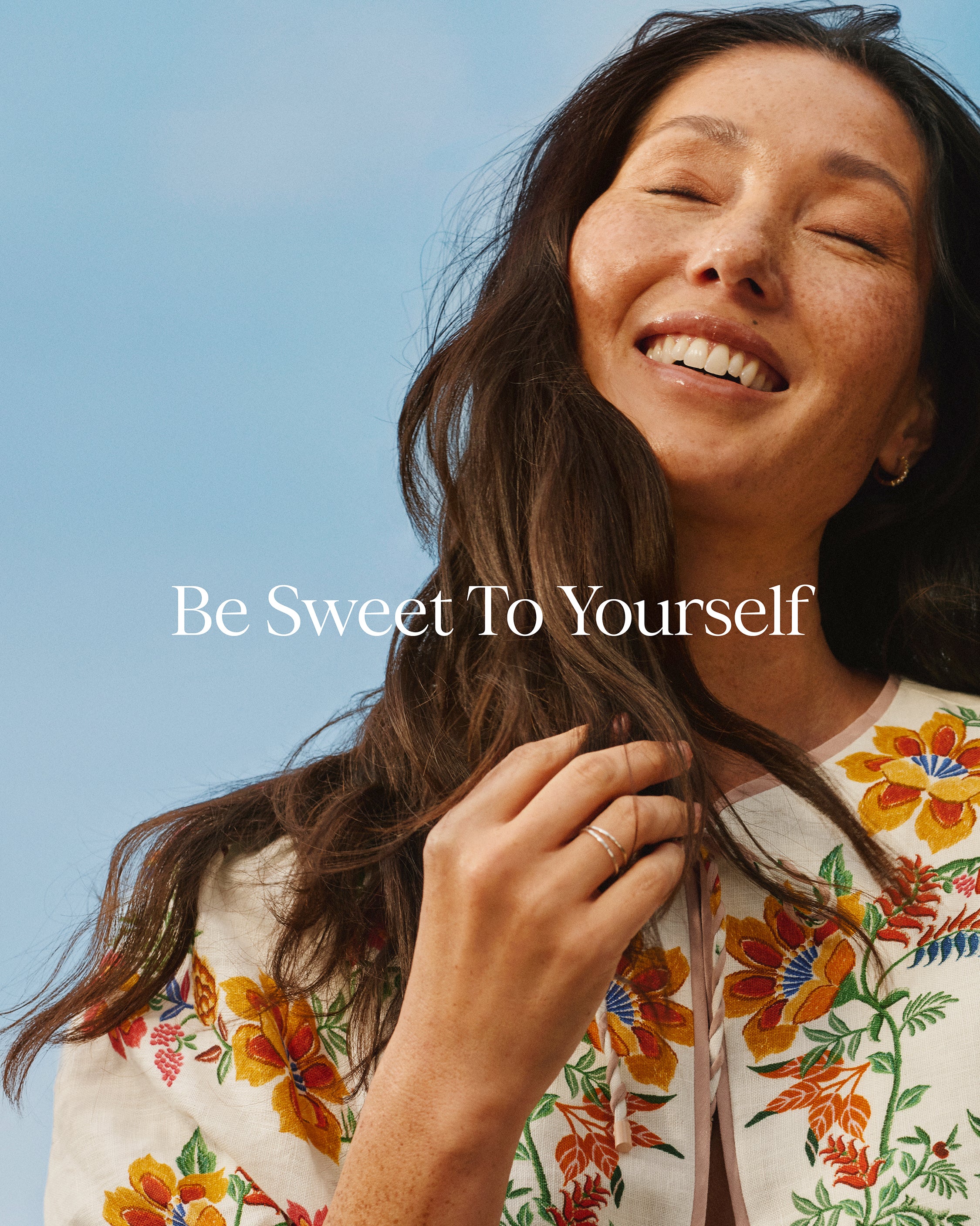 Woman with floral shirt against a blue sky with 'Be Sweet To Yourself' text.