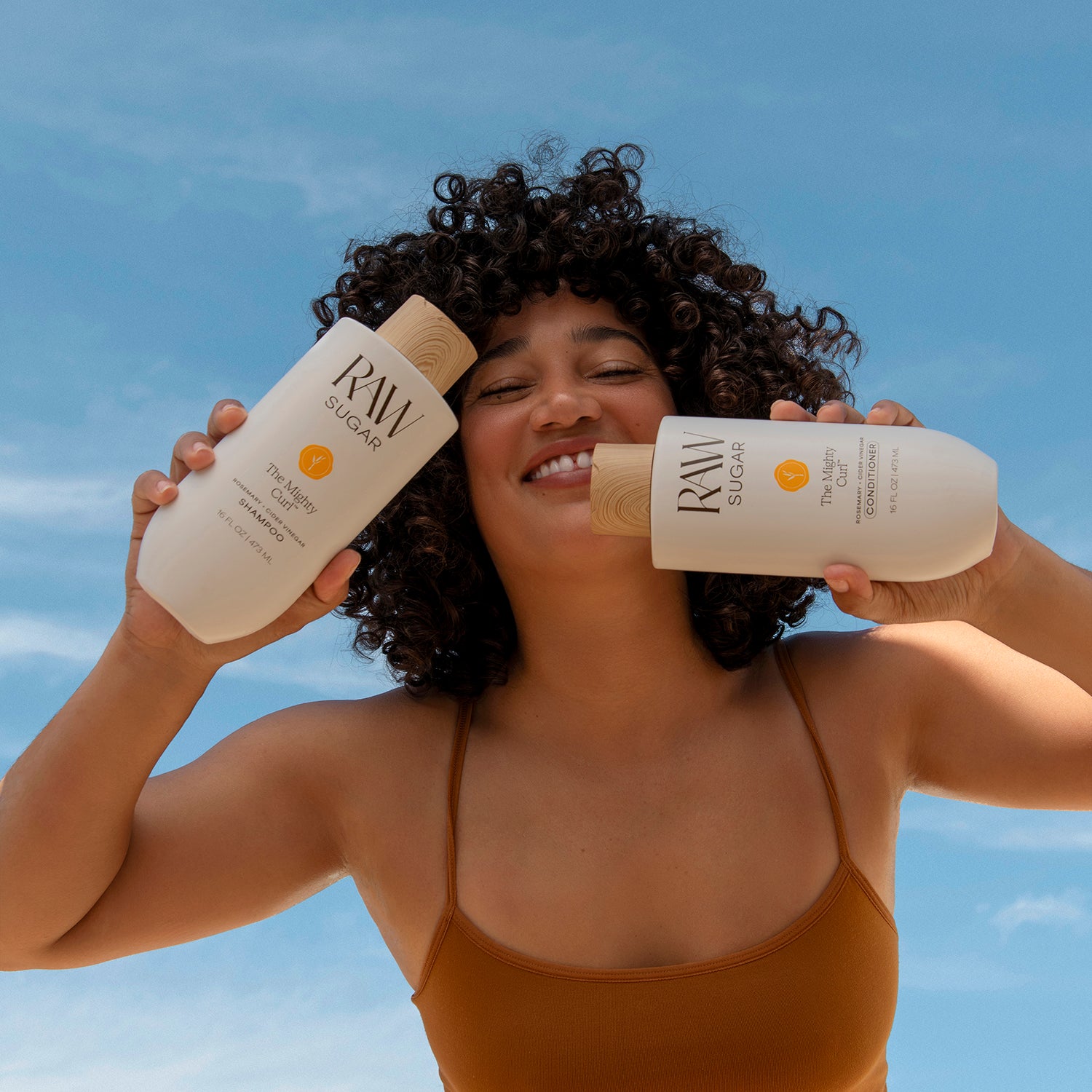 woman smiling and holding Raw Sugar Mighty Curl Shampoo and Conditioner, designed to gently clean curly hair