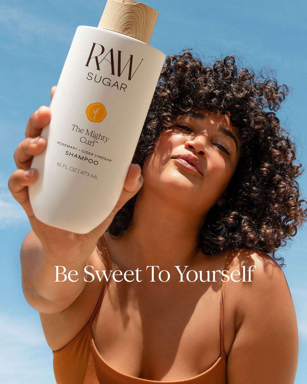 Person holding a bottle of 'Raw Sugar' shampoo against a clear blue sky
