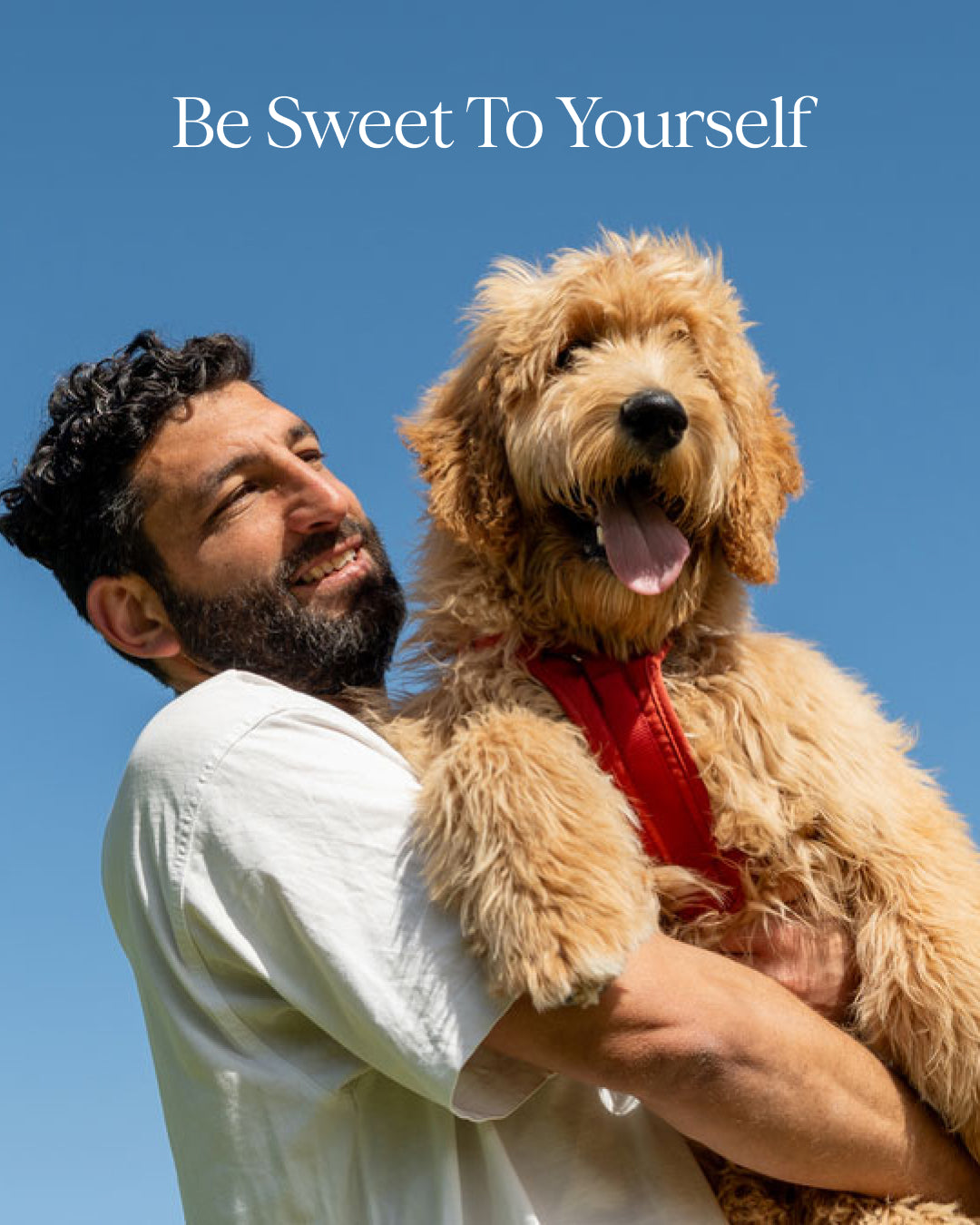 Man holding a fluffy dog with a clear blue sky background and text 'Be Sweet To Yourself'.