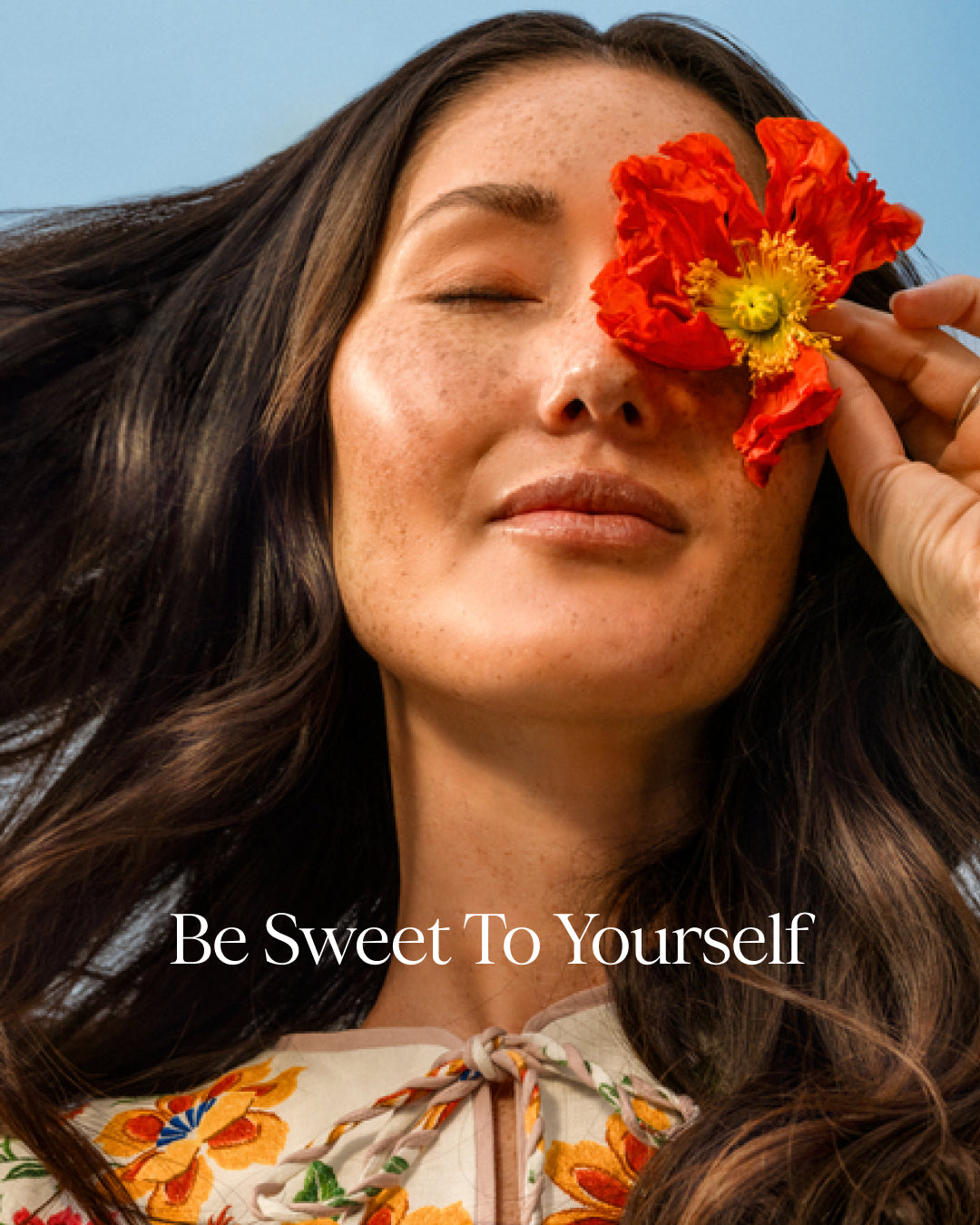 Woman with a red flower covering one eye against a clear blue sky, with 'Be Sweet To Yourself' text.