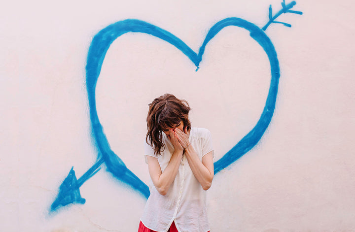 woman smiling into hands in front od heart on pink wall