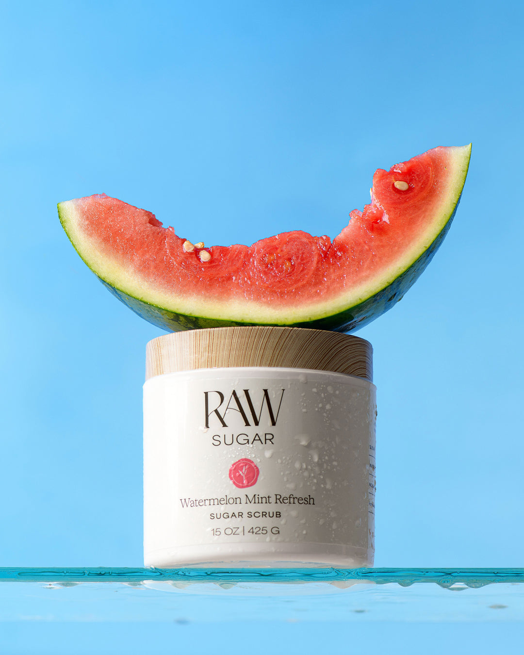 A jar of RAW SUGAR Watermelon Mint Refresh Sugar Scrub sits on a blue reflective surface with a slice of watermelon resting on its lid, against a bright blue background.
