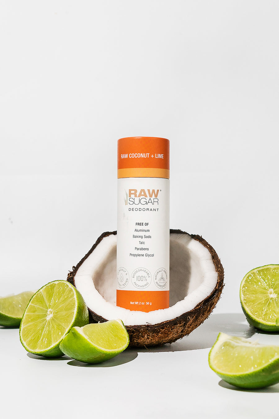 Tube of Raw Coconut + Lime Deodorant sitting in half of a fresh , raw coconut with fresh cut limes scattered around