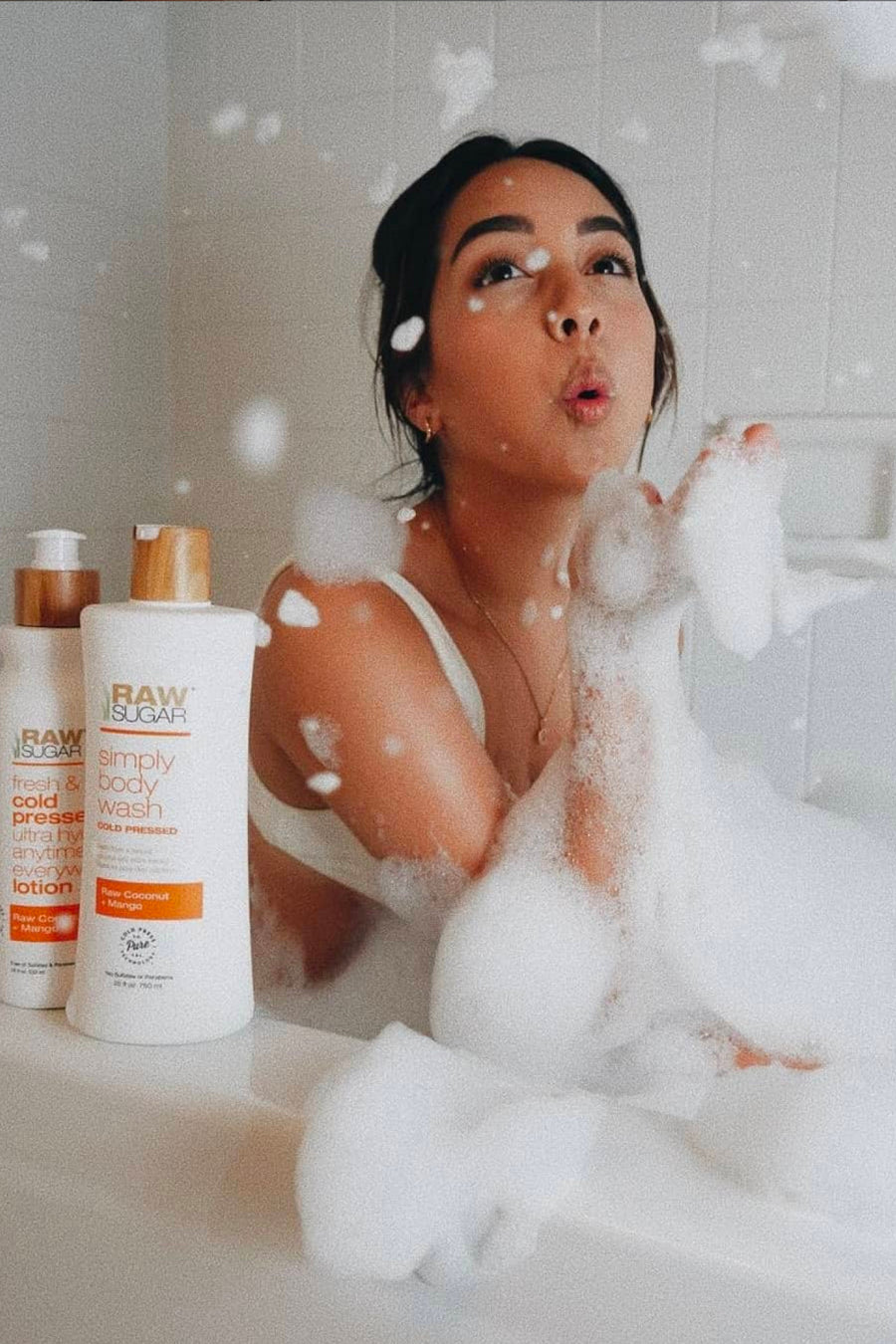 Woman in bathtub playing with bubbles and foam next to Raw Sugar Body Wash and Lotion