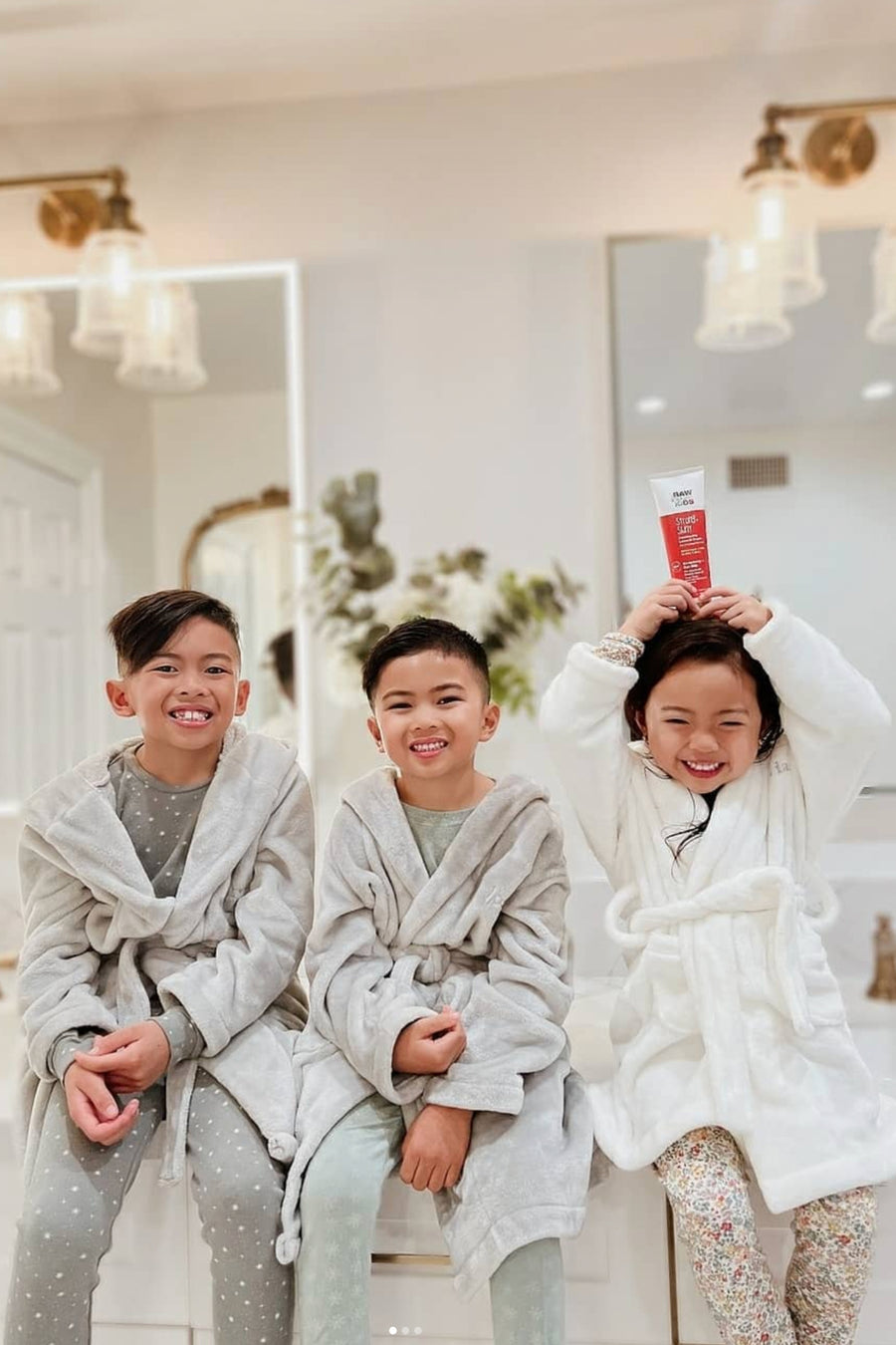 Three Kids in Robes Sitting on a Bathroom Counter with Girl Holding Conditioning Cream