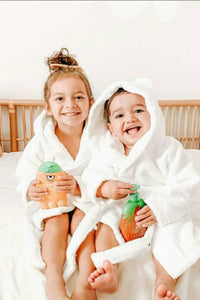 Two Kids in Robes sitting on white bed holding Kids Products