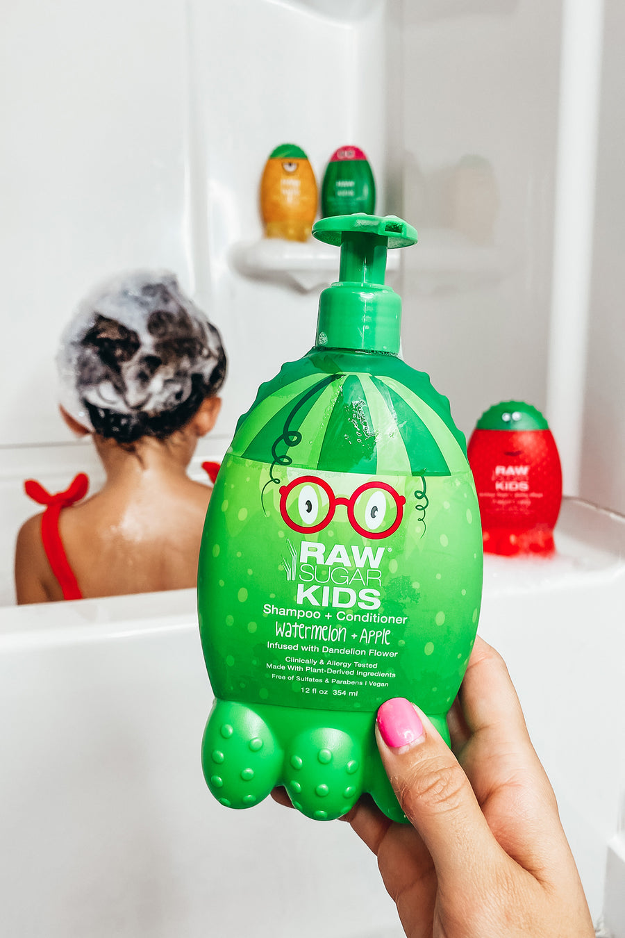 Raw Sugar Kids Watermelon + Apple Shampoo/Conditioner Monster bottle held up in a hand in foreground with little girl with sudsy hair sitting in the bathtub